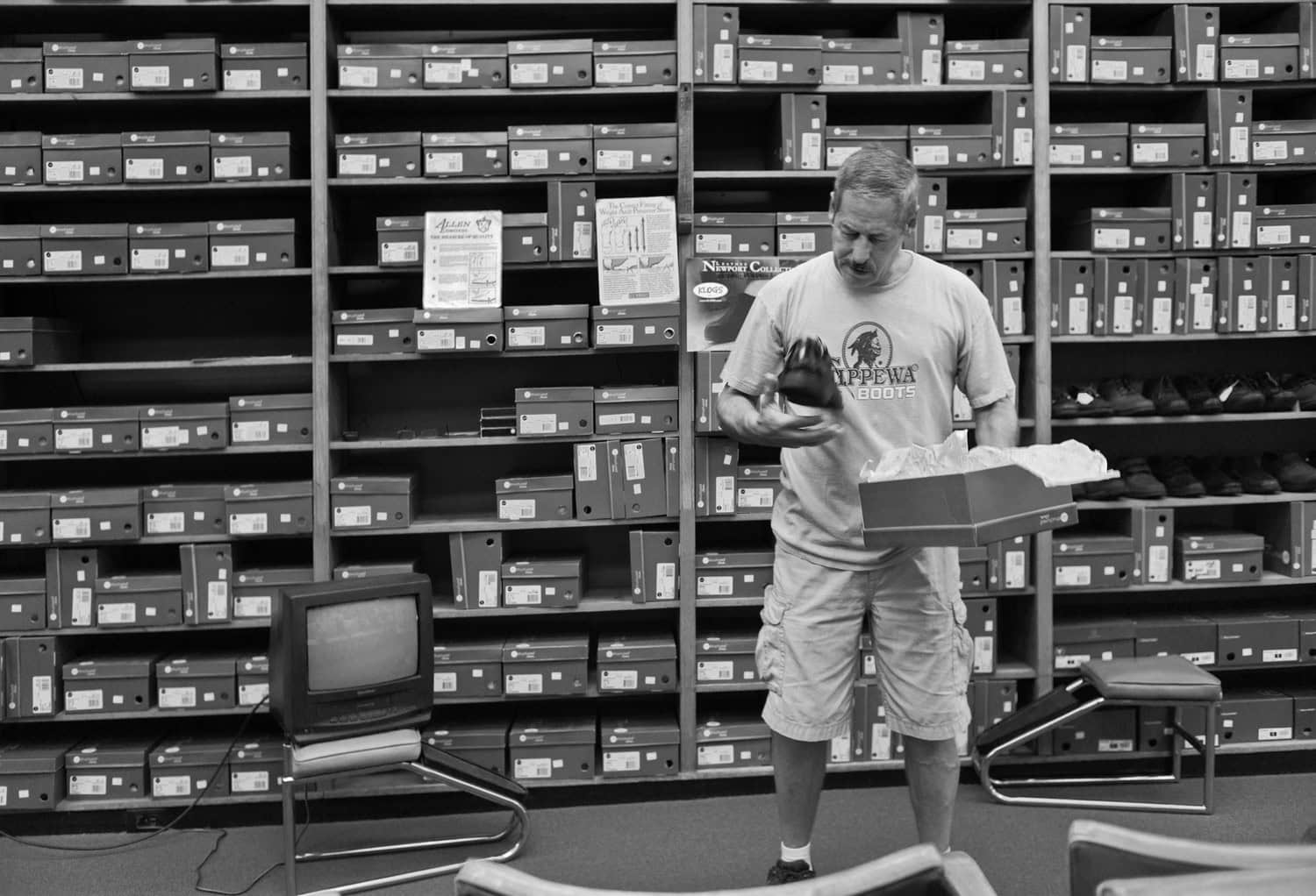 Black-and-white photograph of a man at a shoe store looking at a pair of shoes. Behind him is a wall of shelves with shoe boxes.