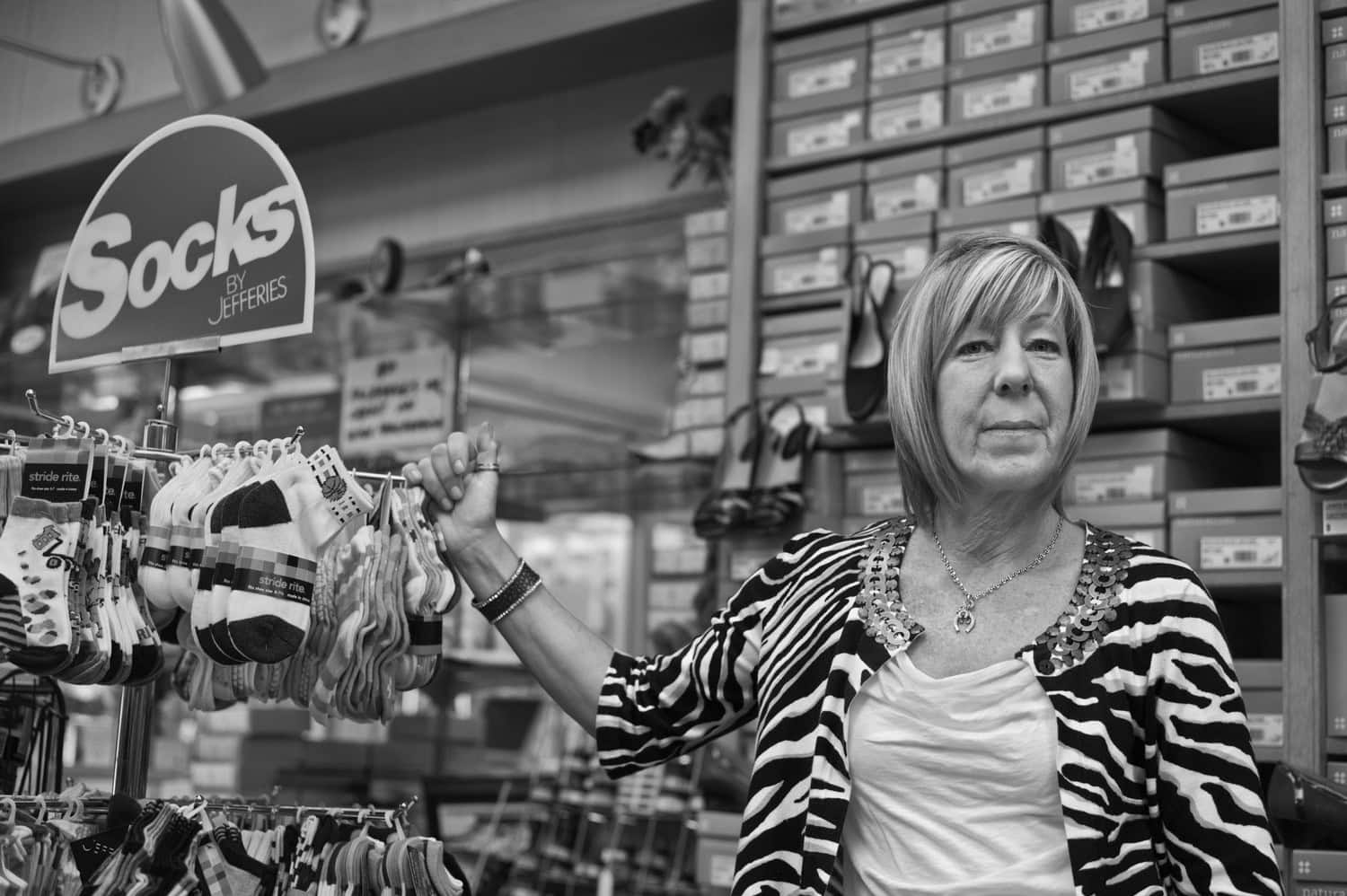 Black-and-white photograph of a middle aged woman with light bobbed hair in a shoe store. Behind her are boxes of shoes and next to her is a small rack with socks. She is wearing a zebra-print jacket and a white t-shirt and is smiling at the camera.