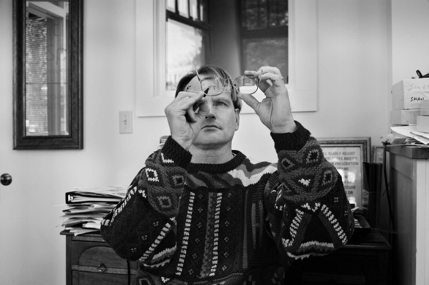 Black-and-white photograph of a man looking into a pair of glasses. He appears to have a desk behind him. He is wearing a sweater and has short dark hair.