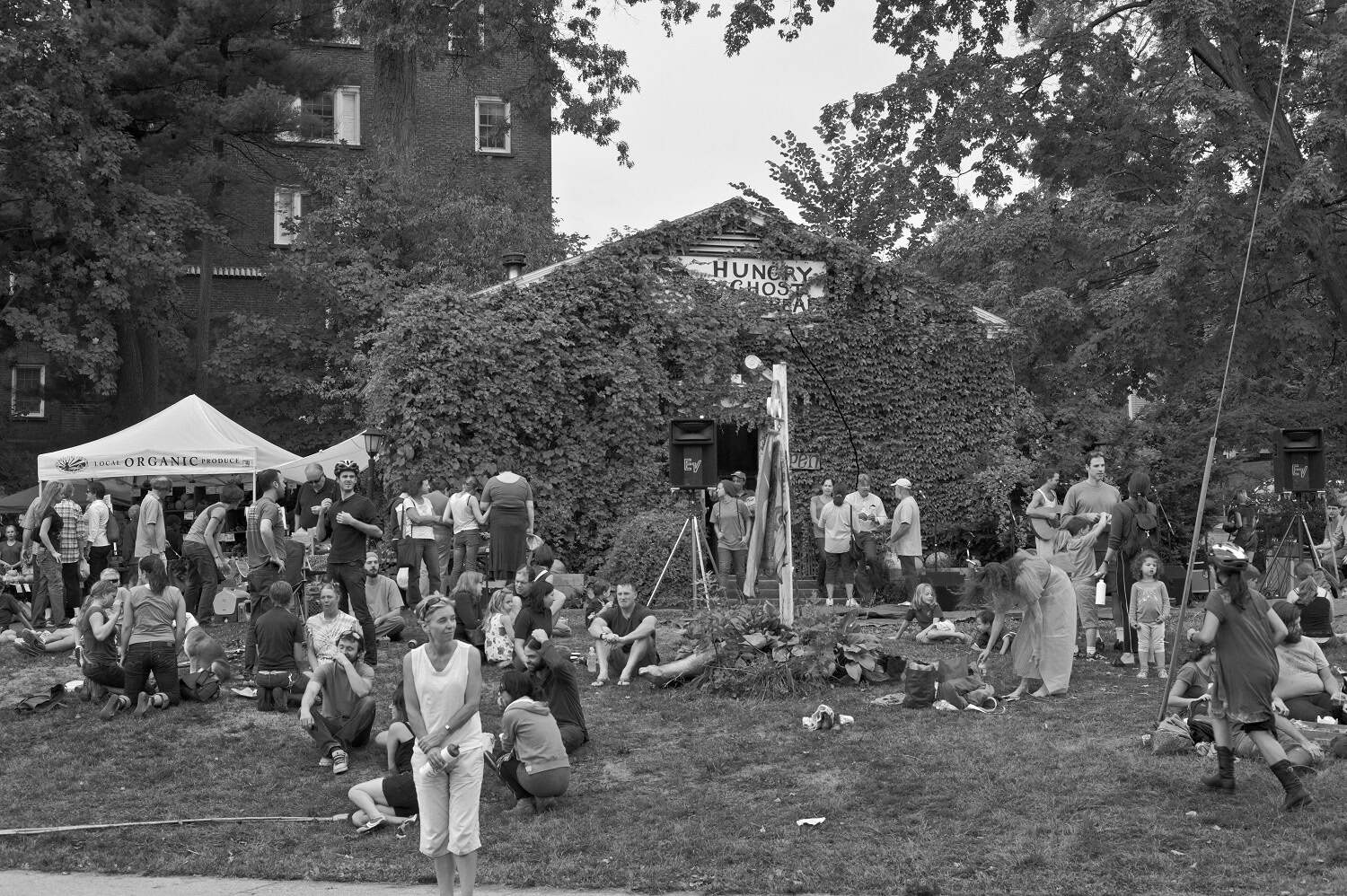Black-and-white photograph of a crowd at Hungry Ghost Bread. The building is covered in ivy, nearly covering the business sign. A tent can be seen in the background with a clutter of people in and around it.