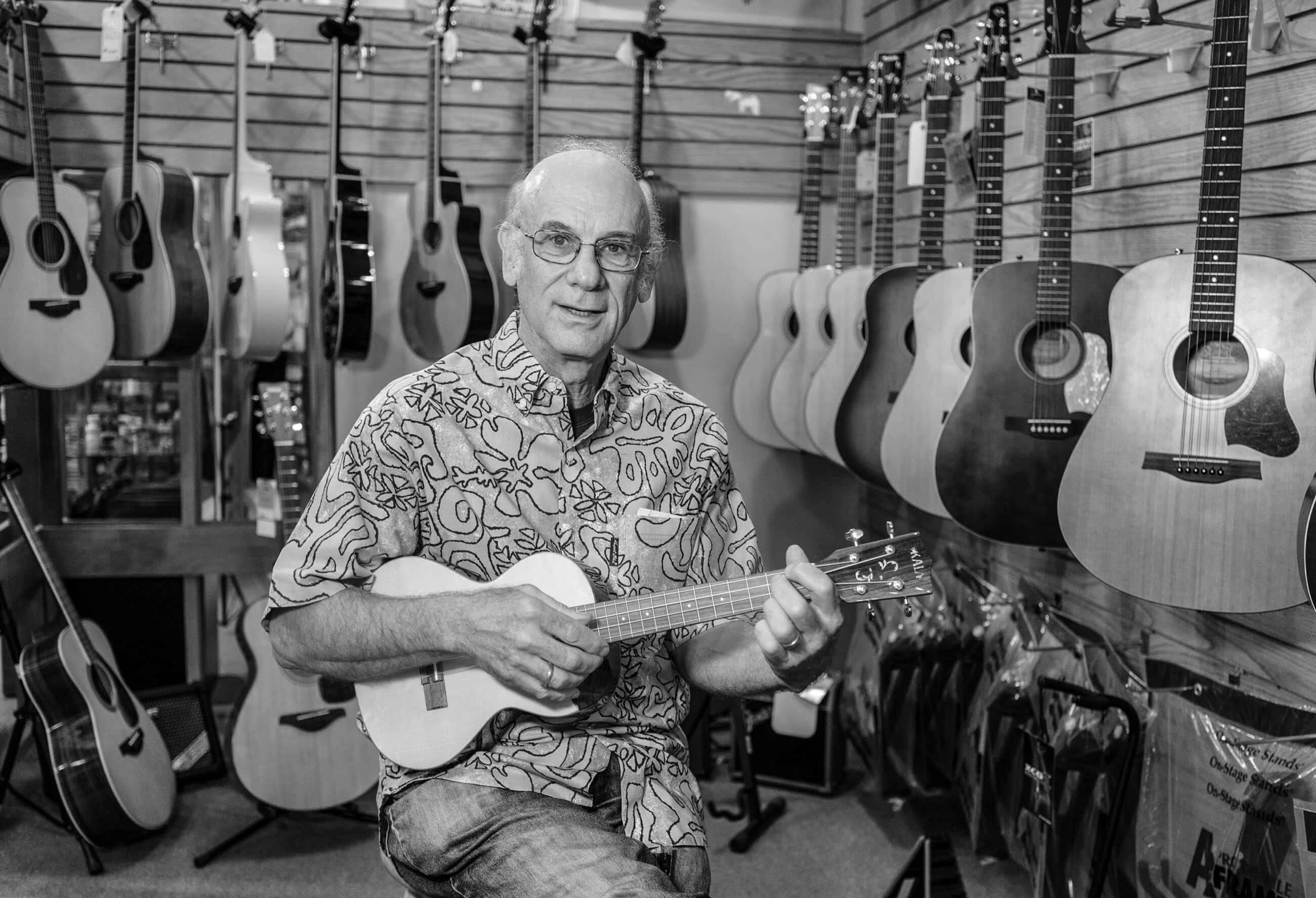 Black-and-white photograph of a matured man with white balding hair and glasses sitting while playing a ukulele or small guitar. Guitars are hanging on the walls around him.