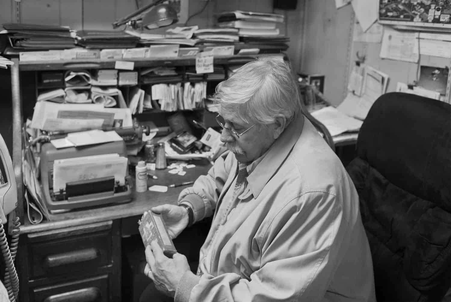Black-and-white photograph of a mature man sitting at his cluttered desk. He has white hair and is wearing a long sleeve button down shirt.