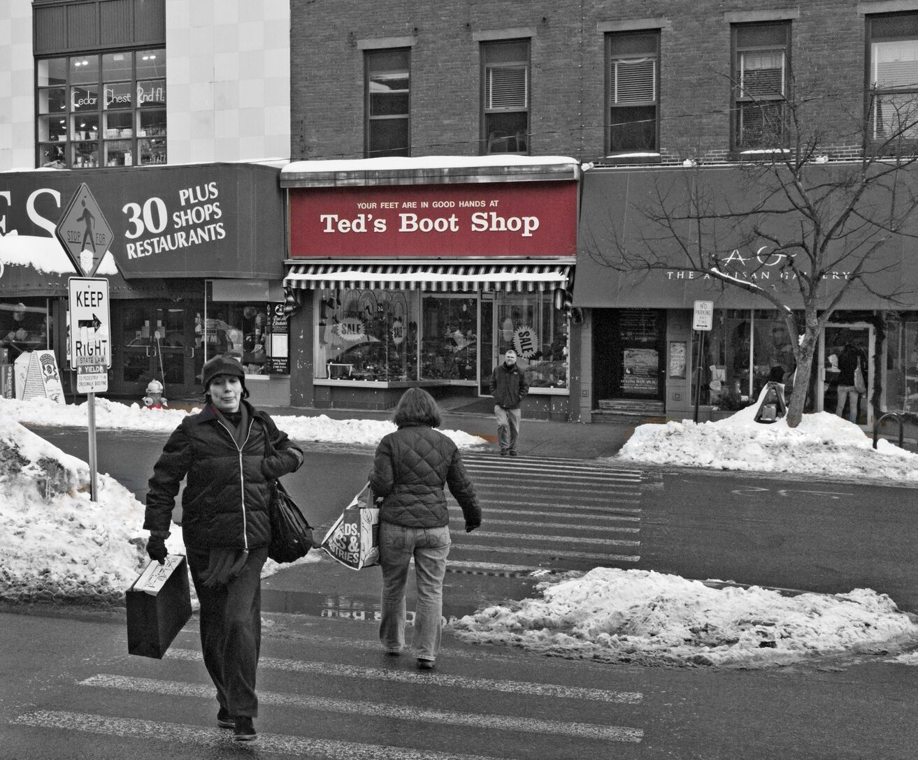 Black-and-white photograph of the crosswalk closest to Thornes Marketplace on Main Street Northampton. The old Ted's Boot Shop (closed) is colored, showing its red storefront sign. People are walking on the crosswalk and snow can be seen melting on the ground.