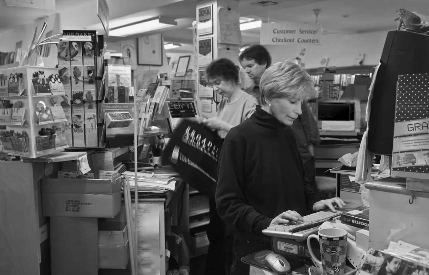 Black-and-white photograph of people working behind the counter at a bookstore. A woman can be seen with short light hair and a turtleneck typing on a computer or cash register. Two other workers are behind her, appearing to be bagging items.