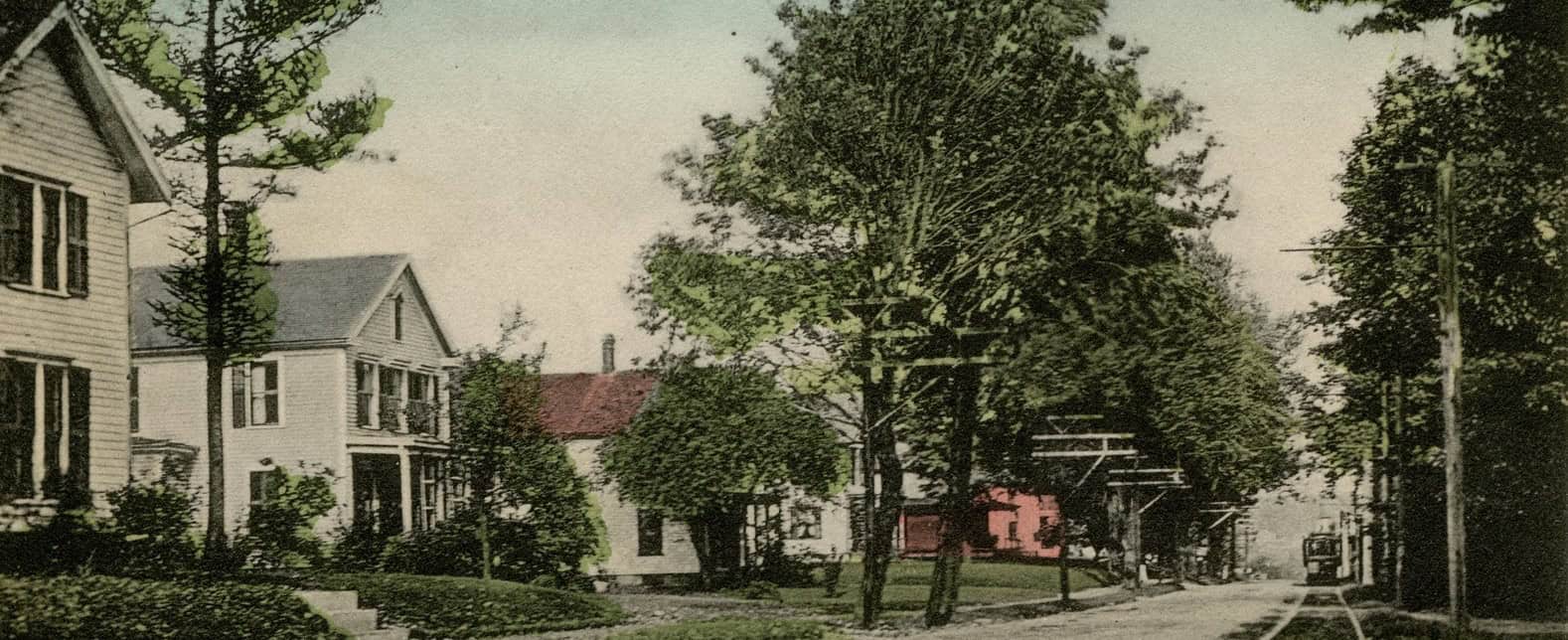 A colorized postcard depicting a view looking down a quiet, tree-lined street. Small, gable-roofed residences border the street. On the left side of the dirt and paved road, a row of two-story wooden houses sits behind manicured green lawns and small stone steps. Lush green trees and utility poles with crossarms line both sides of the street, casting soft shadows across the landscape. In the far distance on the right, a single trolley car rests on a set of tracks that runs parallel to the road.