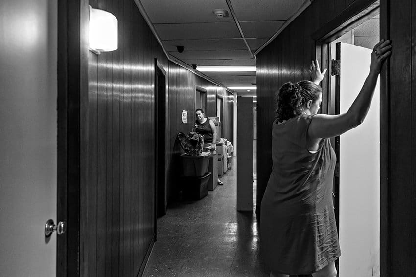 Black-and-white photograph of a woman peering into a room from the hallway with her arms supporting her by holding onto the doorframe. She is wearing a dress and has her black hair tied in a ponytail. In the background a woman can be seen with stacks of moving boxes.