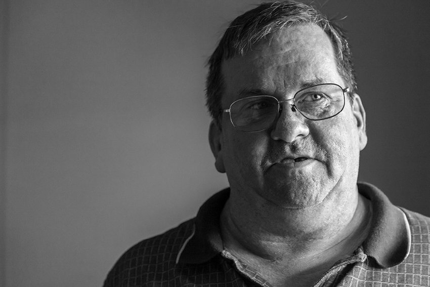 Black-and-white photographic portrait of a middle aged man. He is wearing a dark colored shirt and glasses, and his short graying hair is swept to the side. His face is toward the camera but his eyes are looking to the side, his expression is neutral.