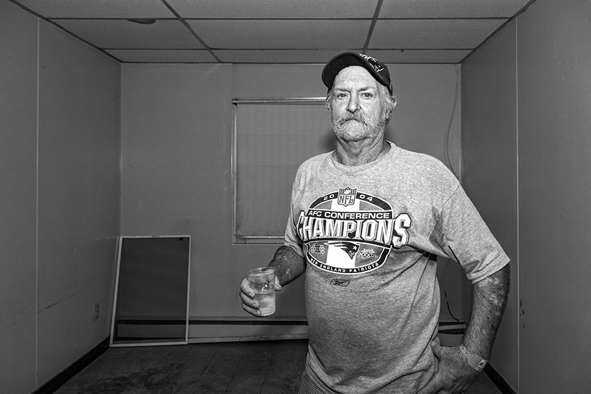 Black-and-white photograph of a man with a Champions shirt on and a baseball cap. The man is matured with graying hair. Behind him is a window screen leaning against the wall and a window with closed blinds. In one of his hands he is holding a plastic cup of water.