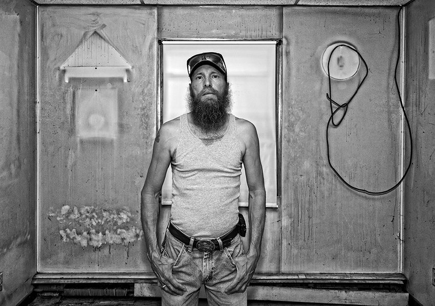 Black-and-white photograph of a middle aged man with a dark long scruffy beard. He is wearing a tank top and jeans and he has a small tattoo on his right shoulder. In the background are smoke-stained walls with white outlines where furniture and wall hangings used to be. A lose cable is hung on the wall.