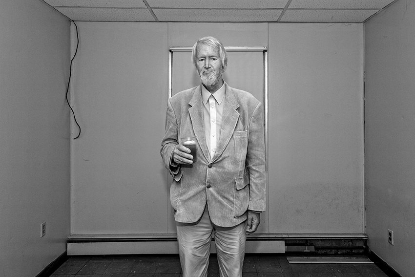 Black-and-white photograph of a matured man standing in the middle of an empty room holding a drink. He has white hair and a white beard and a light colored suit. He is looking toward the camera with a neutral expression.