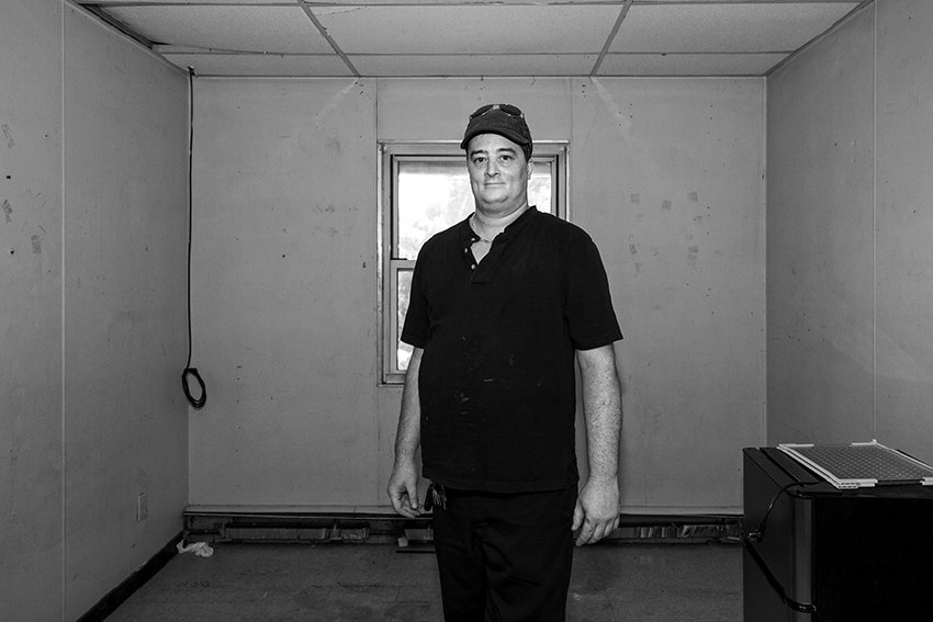 Black-and-white photograph of a middle aged man standing in the middle of an empty room. He is wearing a dark shirt and pants and a baseball hat with sunglasses on his head. He is smiling at the camera.