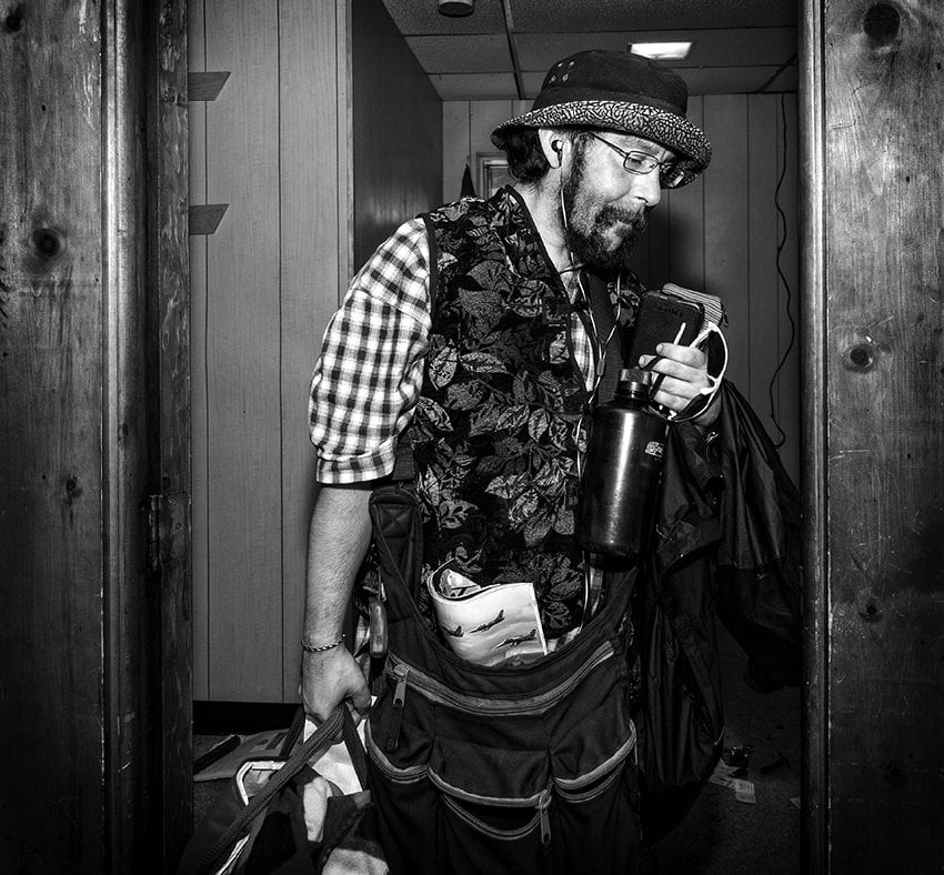 Black-and-white photograph of a man carrying items out of a room. He is wearing a plaid shirt and a vest and has on a hat. He is bearded and wearing glasses.