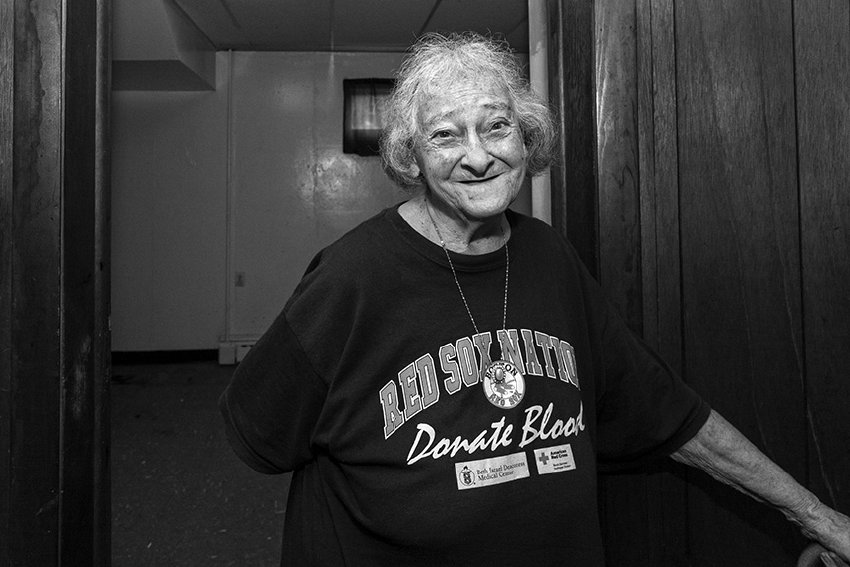 Black-and-white photograph of a matured woman at the entrance way of an empty room. The woman appears to be holding the door or door knob out of the camera's view. She is wearing an oversized Red Sox Donate Blood t-shirt and has white hair. She is smiling toward the camera.