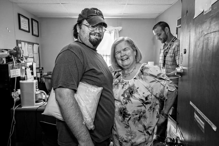 Black-and-white photograph of a man and woman smiling at the camera from the entrance of an open room, with another man facing away in the background. The room is cluttered and appears to be an office space. The man (on the left) is holding a package and the woman on the right has her visible arm on her hip.
