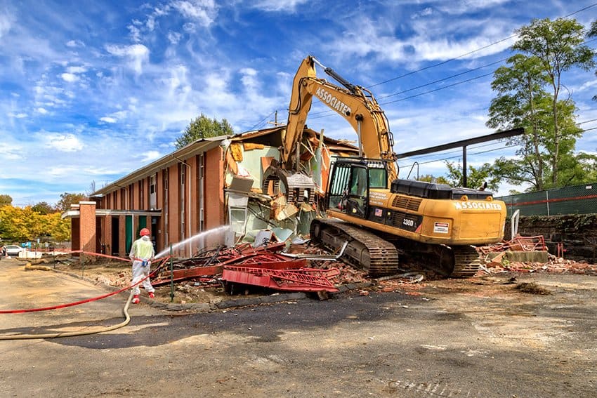 A photograph showing a yellow excavator with "ASSOCIATED" branding demolishing a brick building while a worker hoses down debris.