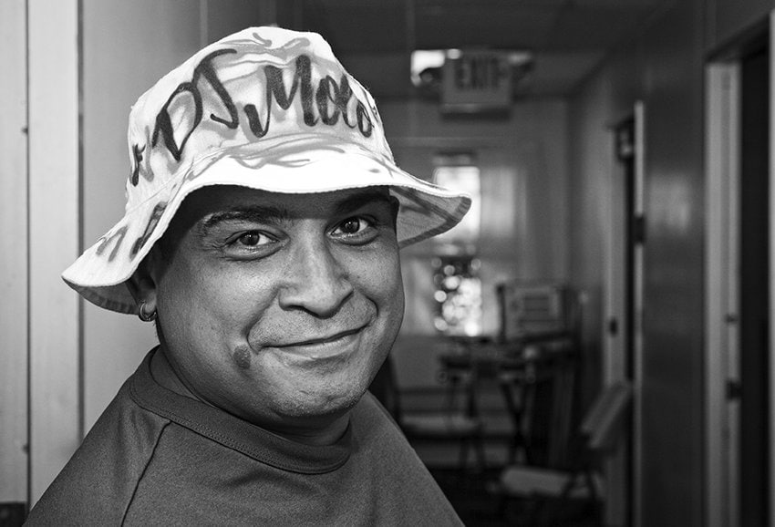 Black-and-white photograph of a man smiling at the camera standing at an open door with a blurred background. He is wearing a hat that has a spray paint inspired design that reads: "DJ MOTO."