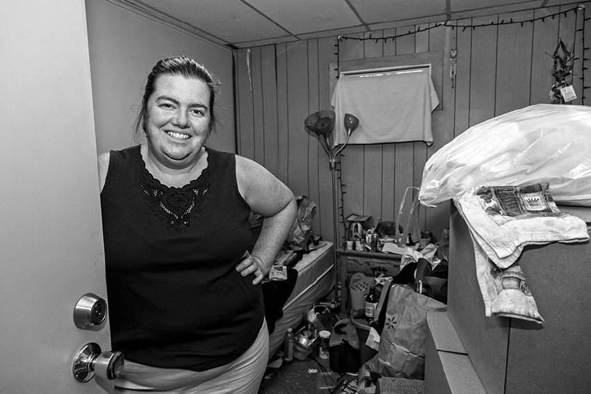 Black-and-white photograph of a woman at the open entrance way of a room. The room is small and cluttered. The woman is wearing a black shirt and her dark hair is pulled away in a ponytail. Her hand is rested on her waist and she is smiling.