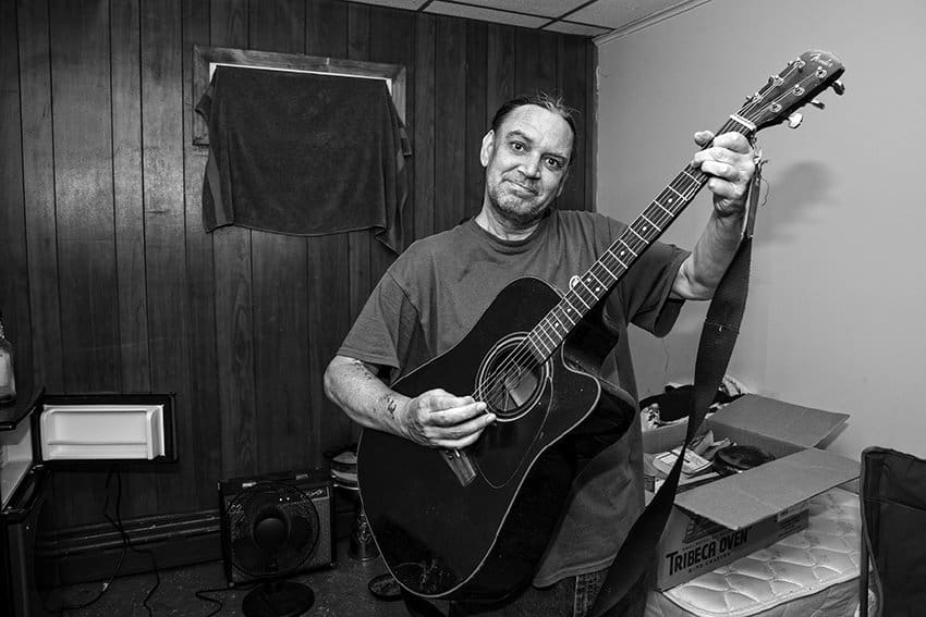Black-and-white photograph of a man holding a black guitar and smiling at the camera. In the background there is a box on a bare mattress, a small fan, a guitar amp, and a window covered by a dark towel. The man is wearing a short sleeve dark top and appears to have his dark hair tied back.