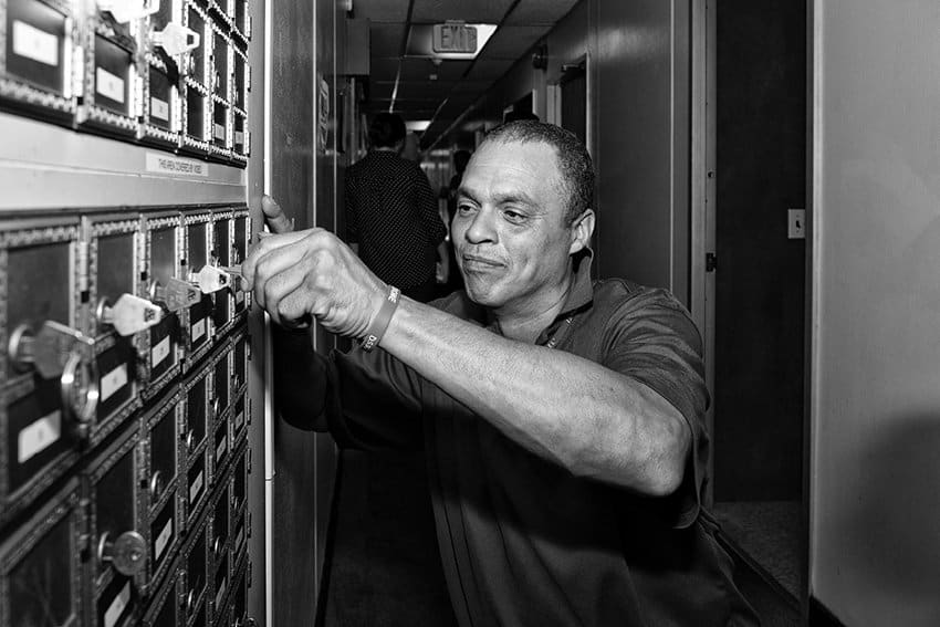 Black-and-white photograph of a man opening a box on the wall. He is looking at the lock and away from the camera. Other figures can be seen in the hallway in the background.