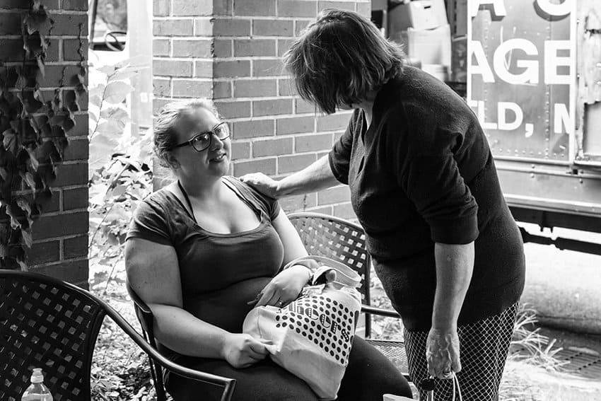 Black-and-white photograph of two women sitting on a porch or under a marquee of a brick building. One woman is sitting on a metal chair with a tote bag on her lap. The other woman is standing and facing the sitting woman, putting her hand on her shoulder. The sitting woman smiles up at her.