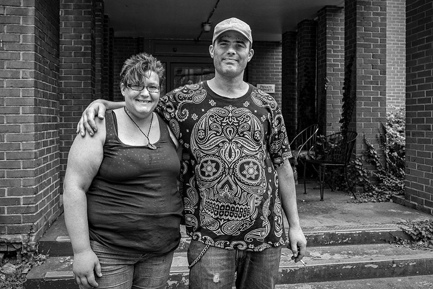 Black-and-white photograph of a couple standing in the outdoor entrance way of a brick building. On the left is a woman with short spikey hair and glasses who is smiling at the camera, wearing a dark tank top and jeans. Next to her is a man with a t-shirt with a Day of the Dead inspired skeleton on it. He is also wearing a baseball cap and jeans. He is smiling and has his arm around the woman.
