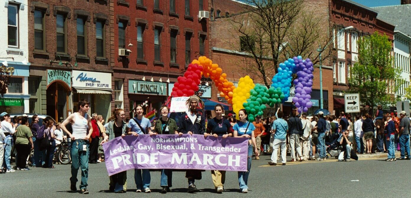 color photograph a parade on Main Street in Northampton. Six people in front are holding a large purple banner reading 'Northamptons Lesbian, Gay, Busexual, & Transgender Pride March.' Behind them are more people marching and a long snaking string of colorful balloons floats above the crowd in rainbow colors. Other people are standingi on either side of the road watching the parade.