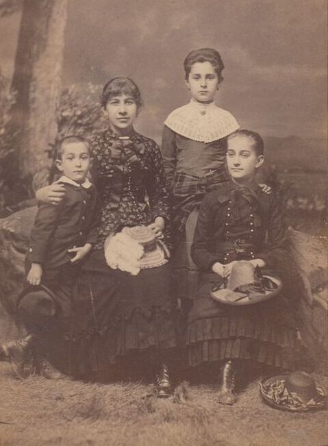 Sepia toned portrait photograph of a woman and three children in a photographer's studio with a painted rural backdrop. The woman is seated with a young boy standing on one sside, and two older girls standing and seated on her other side. All are wearing fancy clothes, and holding their hats, except for one girl's hat which is on the floor.