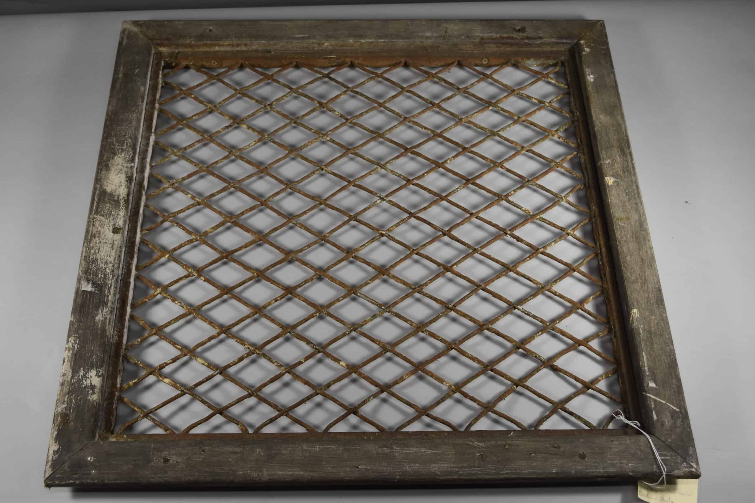 Weathered wooden window grate with a rusty diamond-pattern metal lattice, shown against a plain gray background.