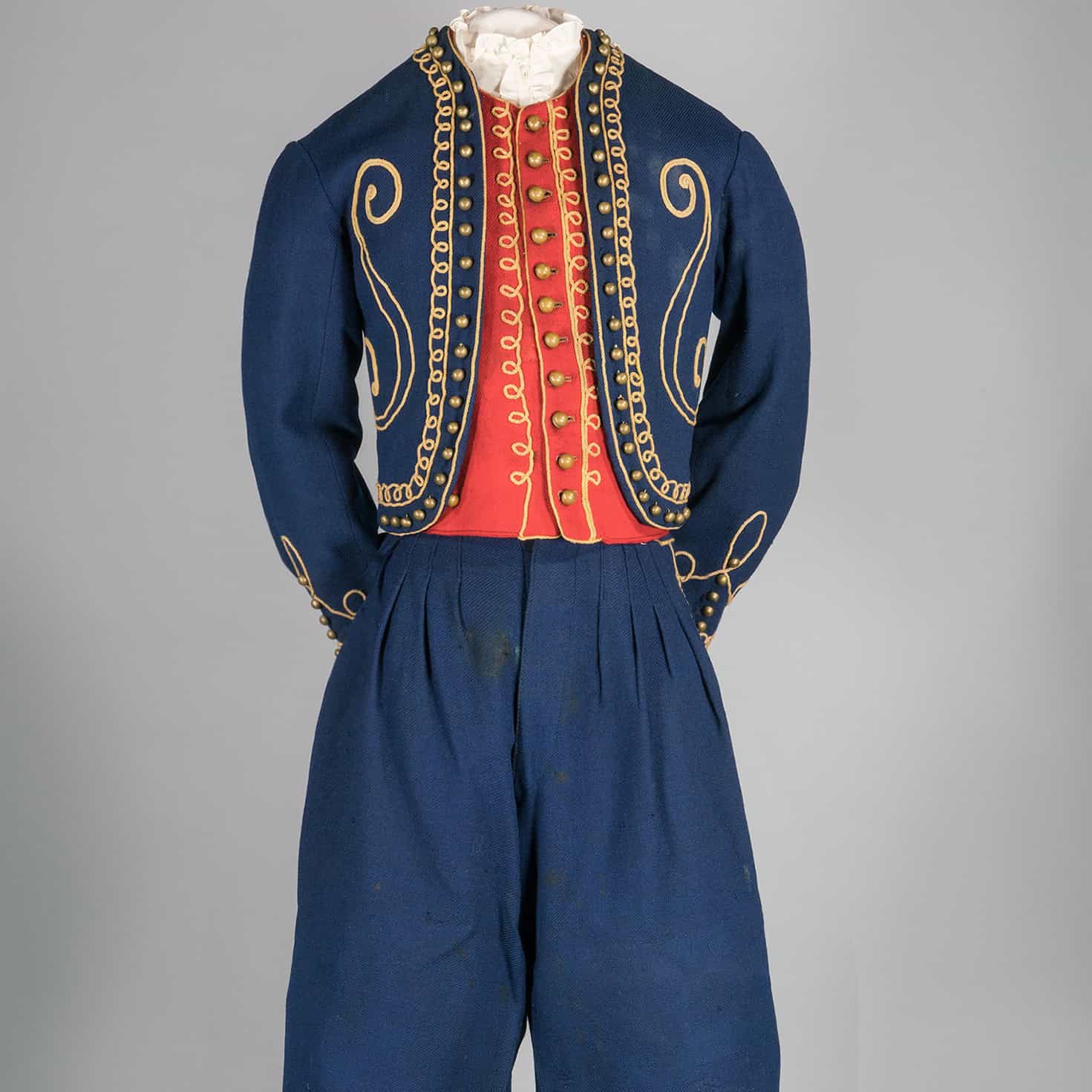 Photograph of a child's costume consisting of a jacket and knickerbocker style pants, both made of dark blue twill wool, and a vest made of red wool twill. All items are embellished with gold cord and brass ball buttons along the edges of jacket, vest and jacket cuffs.