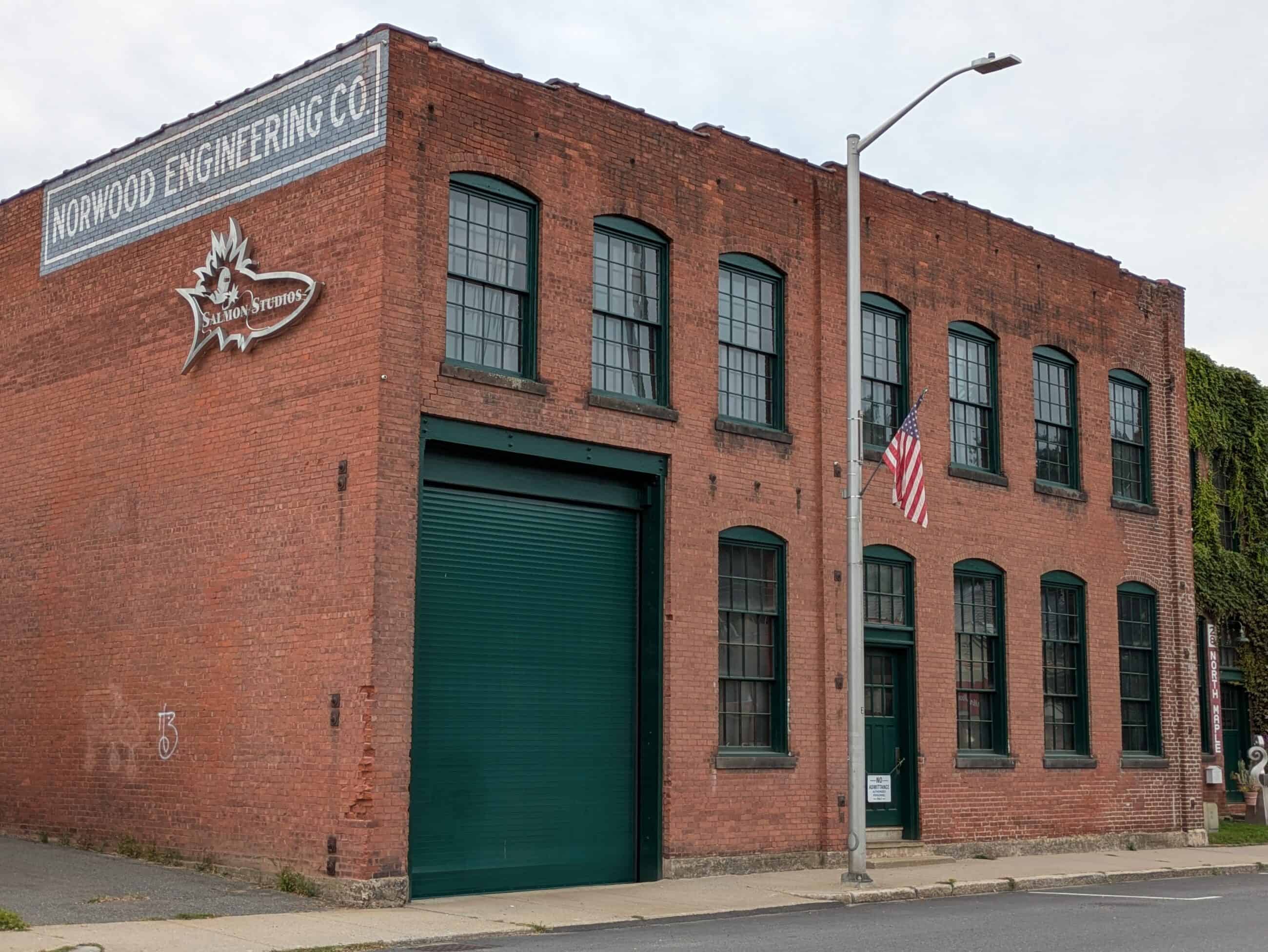 A photograph of the old Norwood Engineering building. The old sign reading: "NORWOOD ENGINEERING CO." can be seen painted on the side. The building is brick with a green garage-style door.