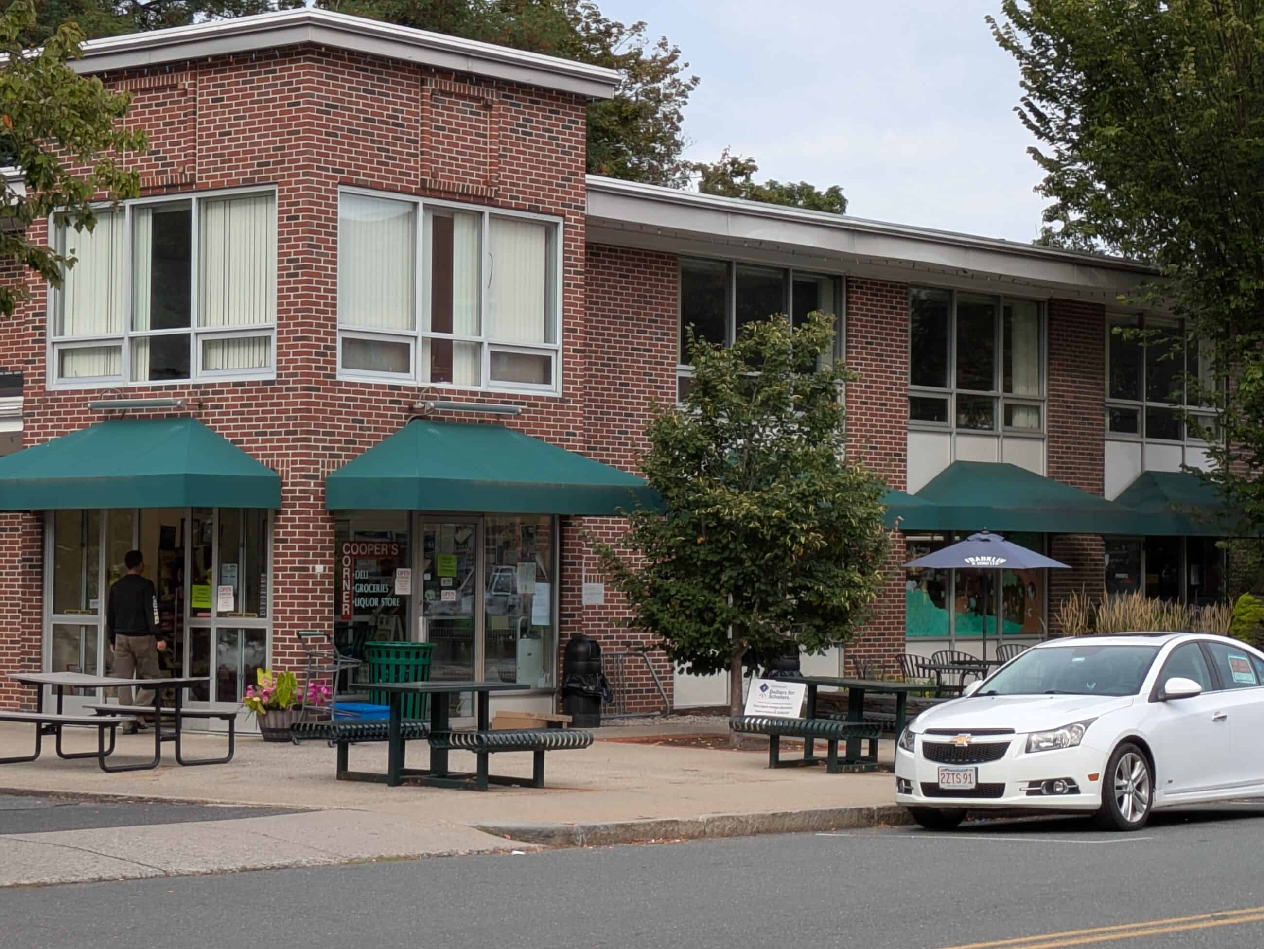 Exterior of red brick building with green marquees. Large windows with white curtains. Picnic tables outside and a white car is parked infront of the store.