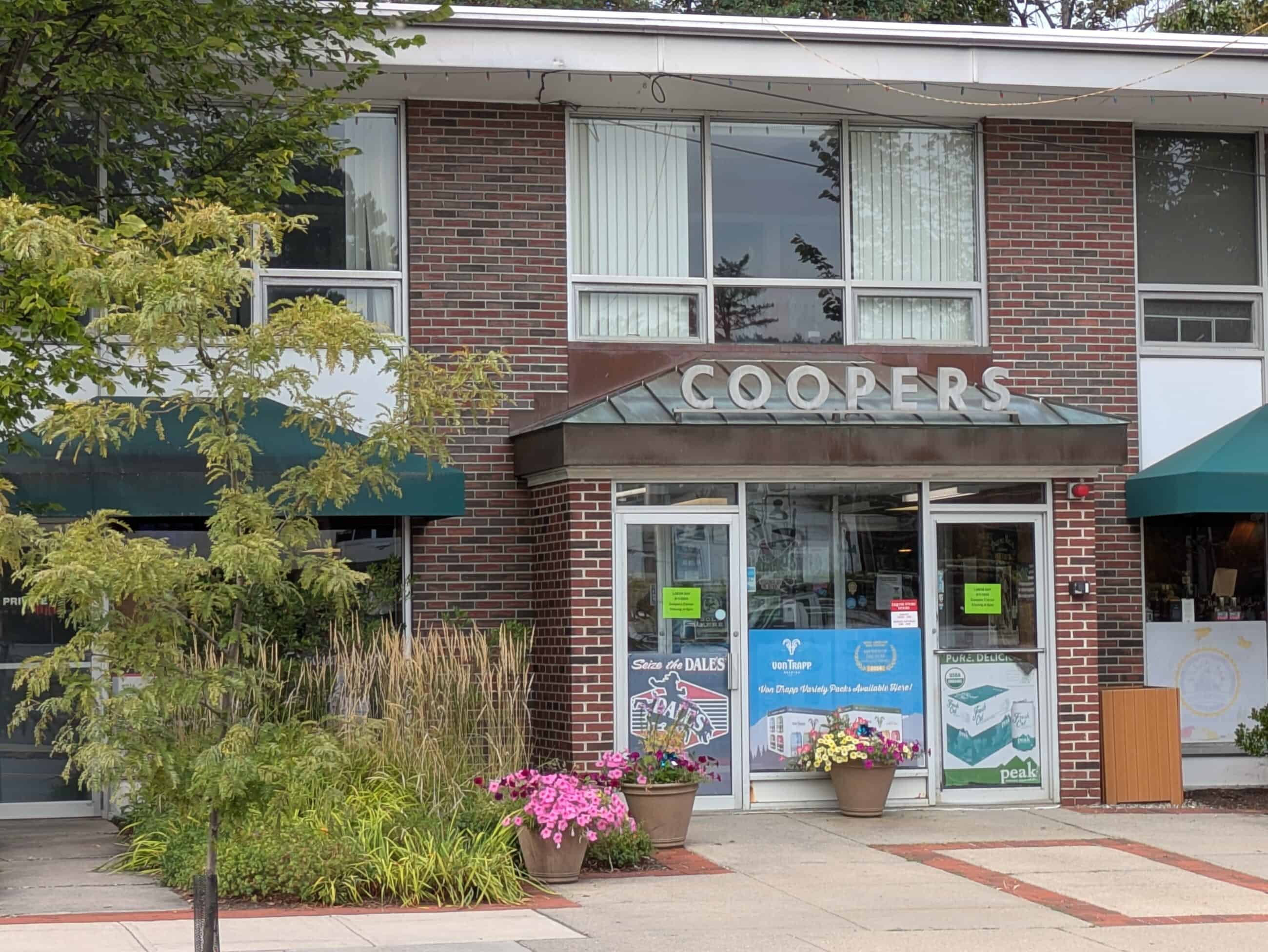 Image of red brick building with green shades and glass entrance with sign that reads "COOPERS." Flowers and greenery infront of building.
