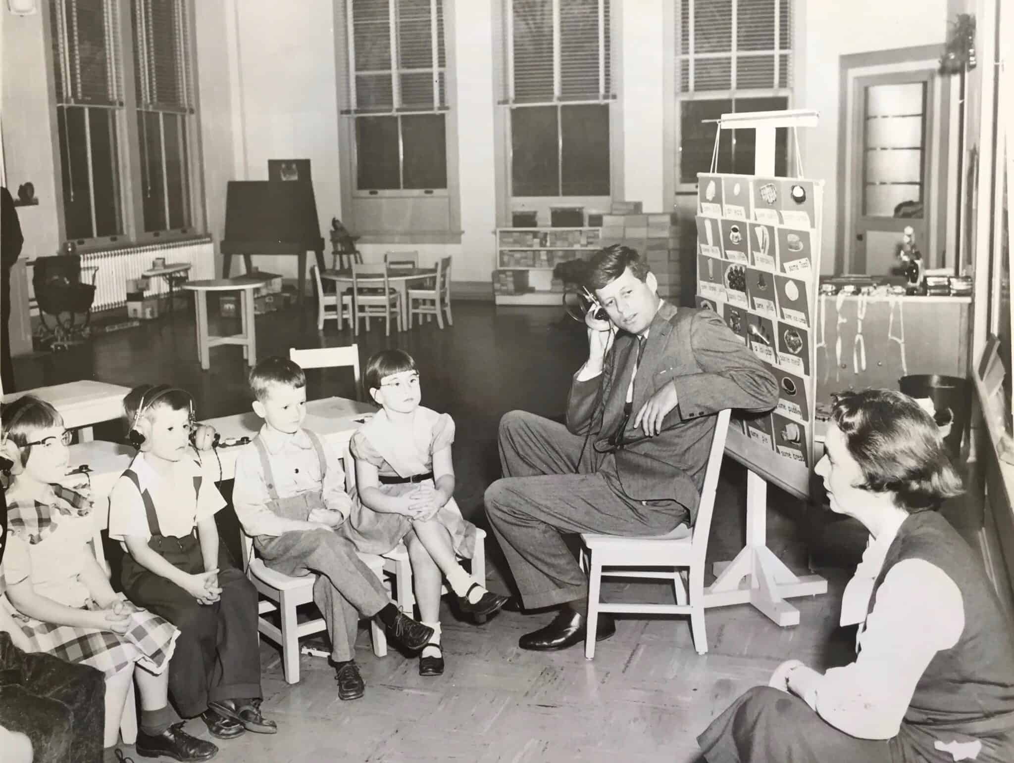 A black-and-white photograph shows a classroom setting where John F. Kennedy, dressed in a dark suit, sits on a small wooden chair while holding a hearing-aid device to his ear. He faces four young children who are seated in a row on small chairs to his left, two of whom are wearing hearing aid devices. A woman sits on the floor to the right, looking toward the group. Behind Kennedy stands a tall, vertical flip chart displaying rows of simple illustrations and text. The classroom is spacious, featuring large windows with blinds, a student desk and chair set in the foreground, and additional small tables and chairs arranged in the background.