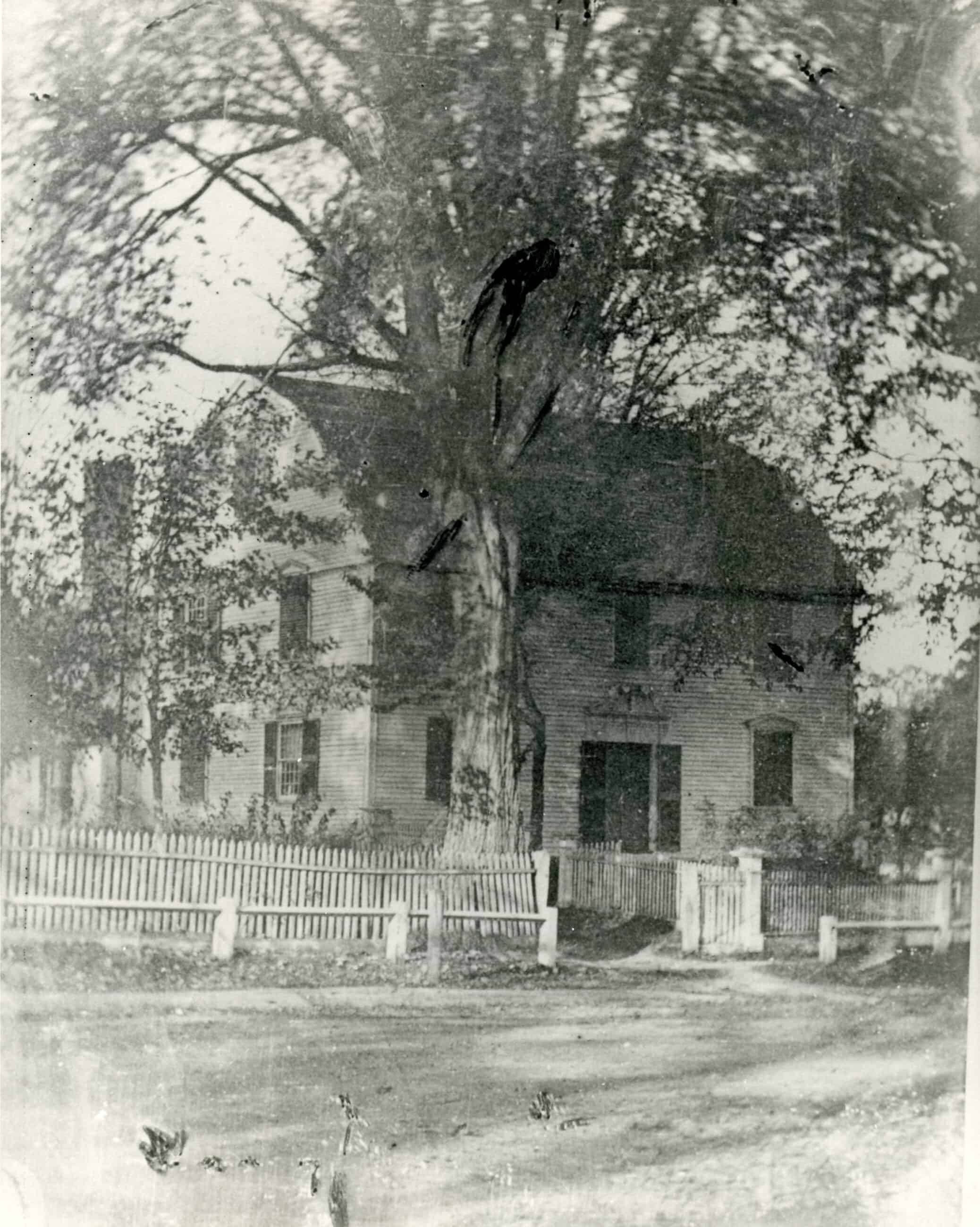 Black-and-white photograph of a house with a half-oval roof. A white picket fence with an open gate surrounds the house.
