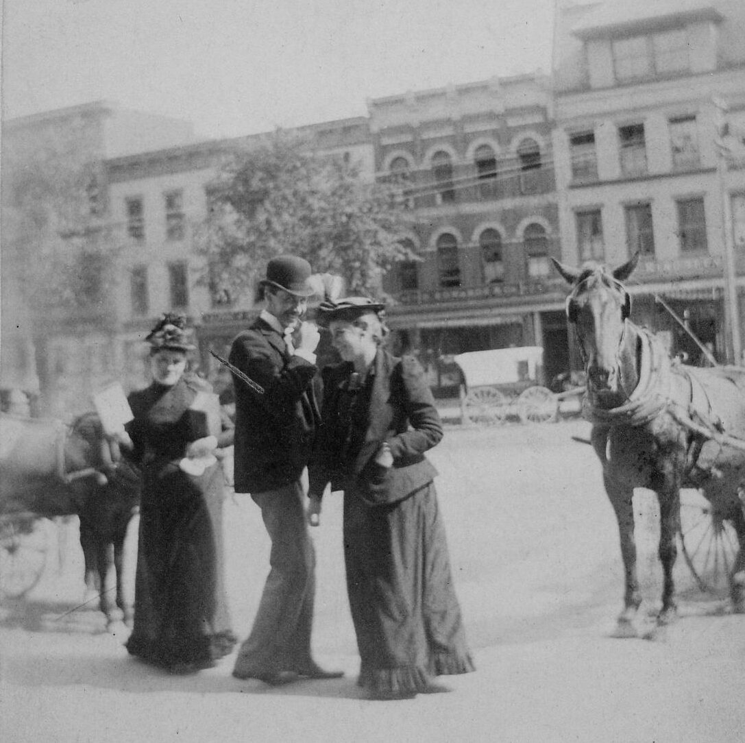 Black-and-white photograph of three people on Main Street. One man wearing a hat and suit is speaking to a woman in a black dress and suit jacket and they are laughing together. A horse can be seen peaking out the side of camera's view. Another woman stands in the background facing the camera. Another horse stands behind her and one wheel of the carriage the horse is tied to can be seen.