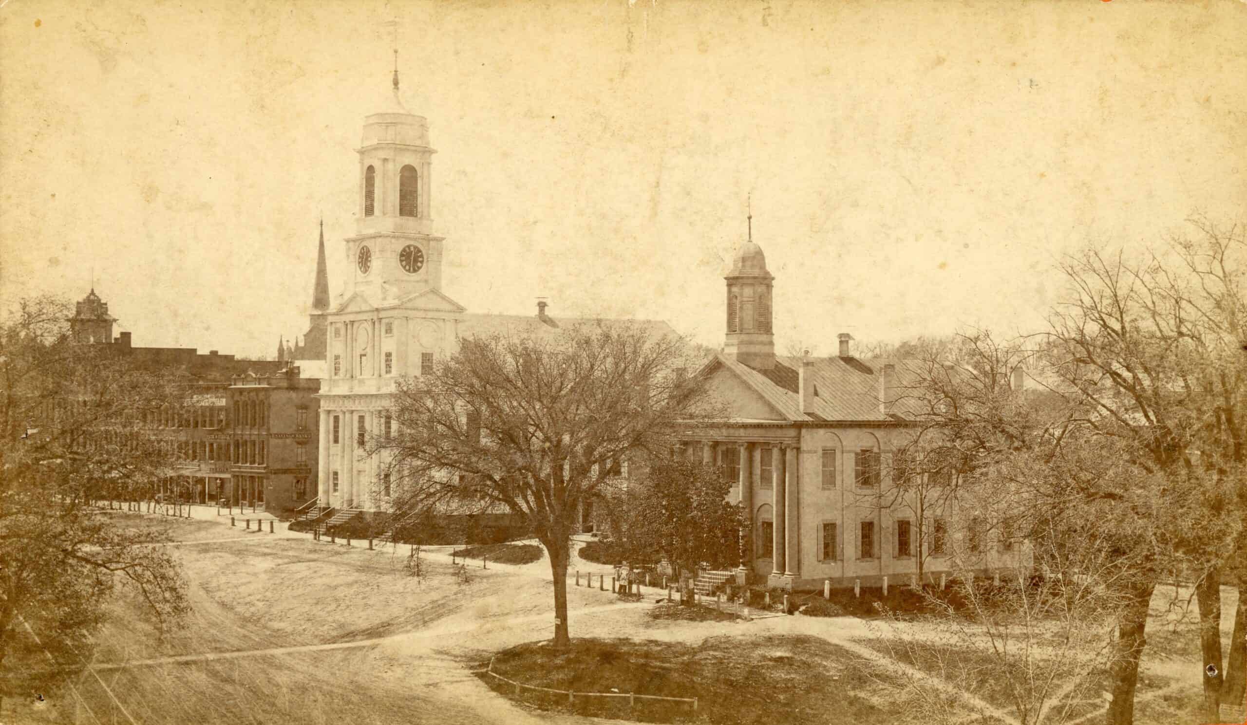 Black-and-white photograph of the old Church Meetinghouse and court house on Main Street. The dirt road infront can be seen, alongside nature and trees lining the Main Street.
