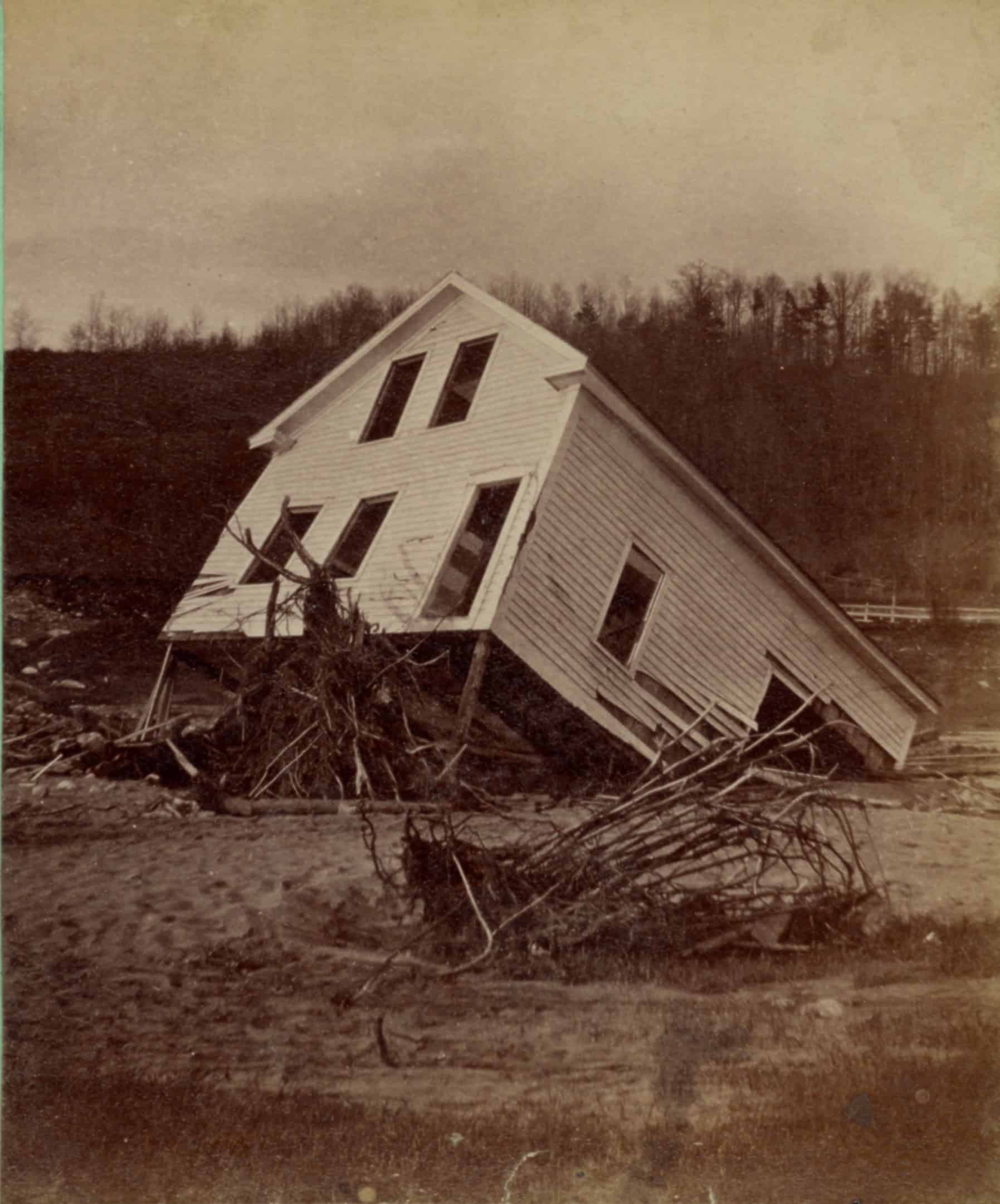 Image of a white house tipped on one end and partially destroyed by flood waters