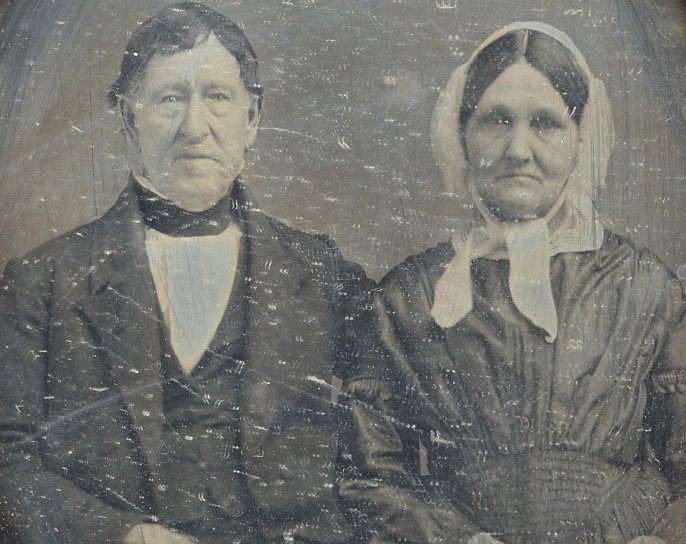 Black-and-white portrait of Isaac Damon and his wife Sophia Strong Damon. The couple are sitting next to eachother with rested faces. Isaac Damon wears a black-and-white suit and Sophia Strong Damon wears a simple black dress and a white head covering tied in the front in a loose bow.