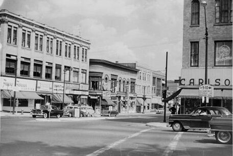Black-and-white photograph captures a mid-20th-century street intersection in a downtown commercial district. On the left side of the street, a long row of contiguous brick commercial buildings stretches into the background, featuring multi-story facades with dense rows of rectangular windows, ground-floor storefronts with fabric awnings, and various shop signs. Several classic 1950s-era automobiles are parked parallel along the left curb. The street itself is wide, marked with white crosswalk lines in the foreground, and leads toward a distant bend under a cloudy, bright sky. On the right foreground, the corner of a prominent multi-story brick building stands tall, featuring a large storefront sign that reads "CARLSO..." in bold block lettering, an awning, and a window advertisement displaying a circular emblem with the word "LOANS". A dark classic sedan is captured in motion, turning through the intersection in front of this corner building, while a tall street lamp and a traffic sign marking routes "9-10" stand nearby on the sidewalk.