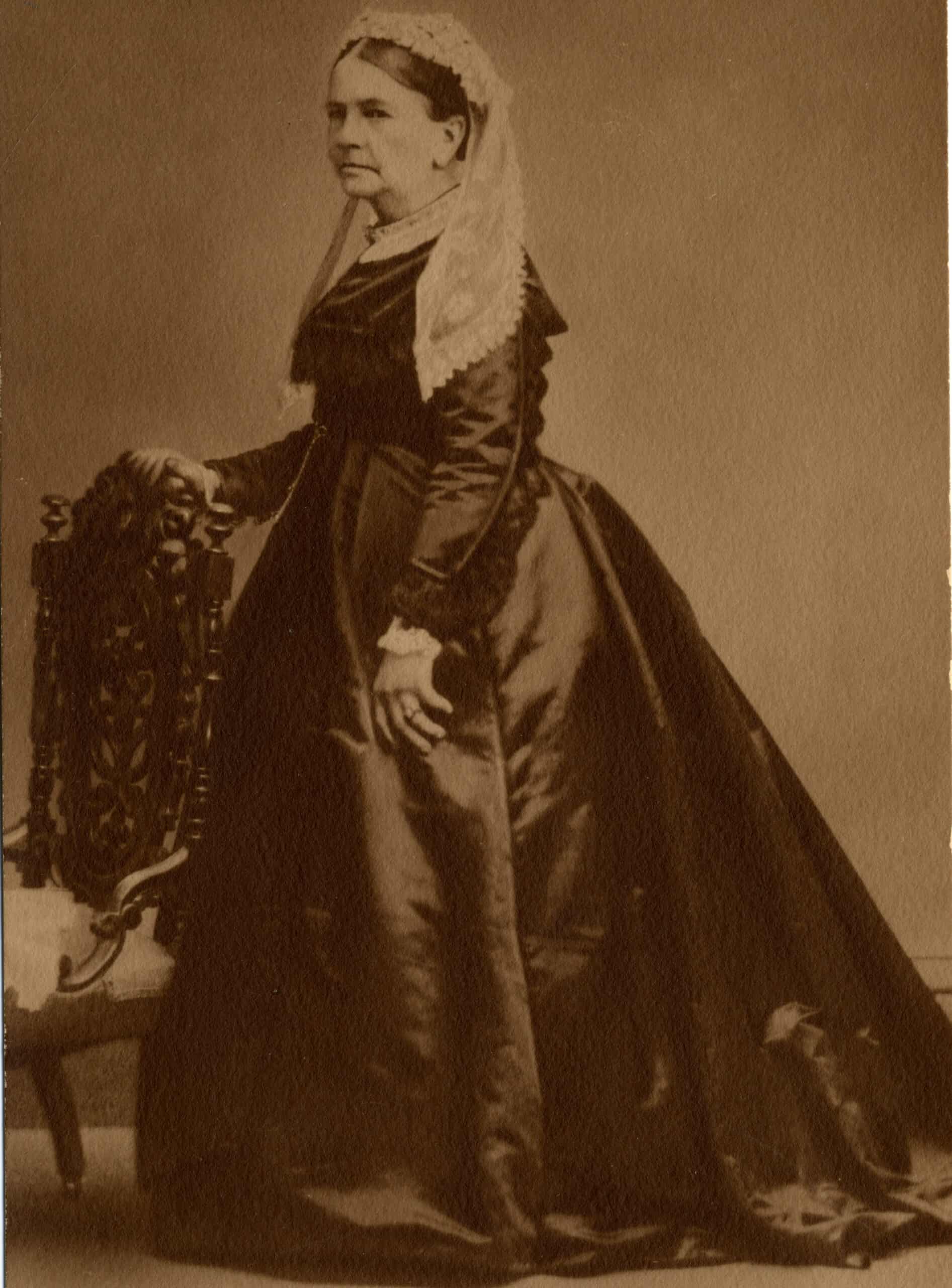 Full-length black-and-white photograph of a mature woman standing in profile, facing left, with her right hand resting on the back of an ornate wooden chair. She has dark hair styled close to her head, topped with a delicate, white lace head covering that drapes over her shoulders. She wears a dark, long-sleeved dress with a high collar, trimmed with lace at the neck and cuffs. The voluminous skirt gathers at the waist and flows down to the floor, pooling slightly at her feet. The background is a solid, neutral color, and the overall image has a textured, matte finish.