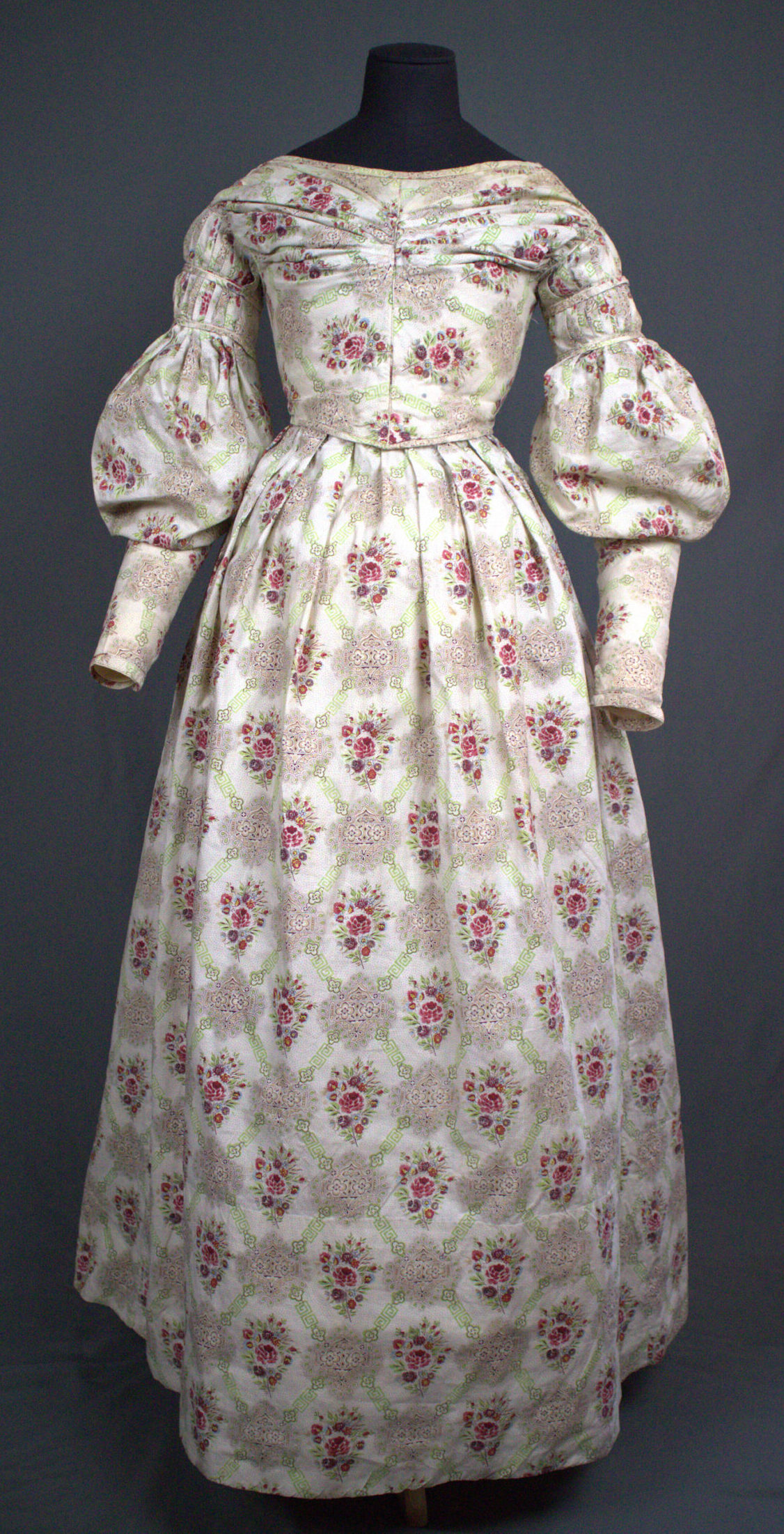 cotton dress with a pattern of pink roses within a gridded design featuring gothic medallion motifs. The sleeves are pleated at the shoulder with the fullness controlled around the upper arm with narrow bands of self fabric. The waistline is a shallow V, The neckline is wide and round. The bodice is gathered across the center of the bustline to the shoulders. The skirt is pleated all around the waist.