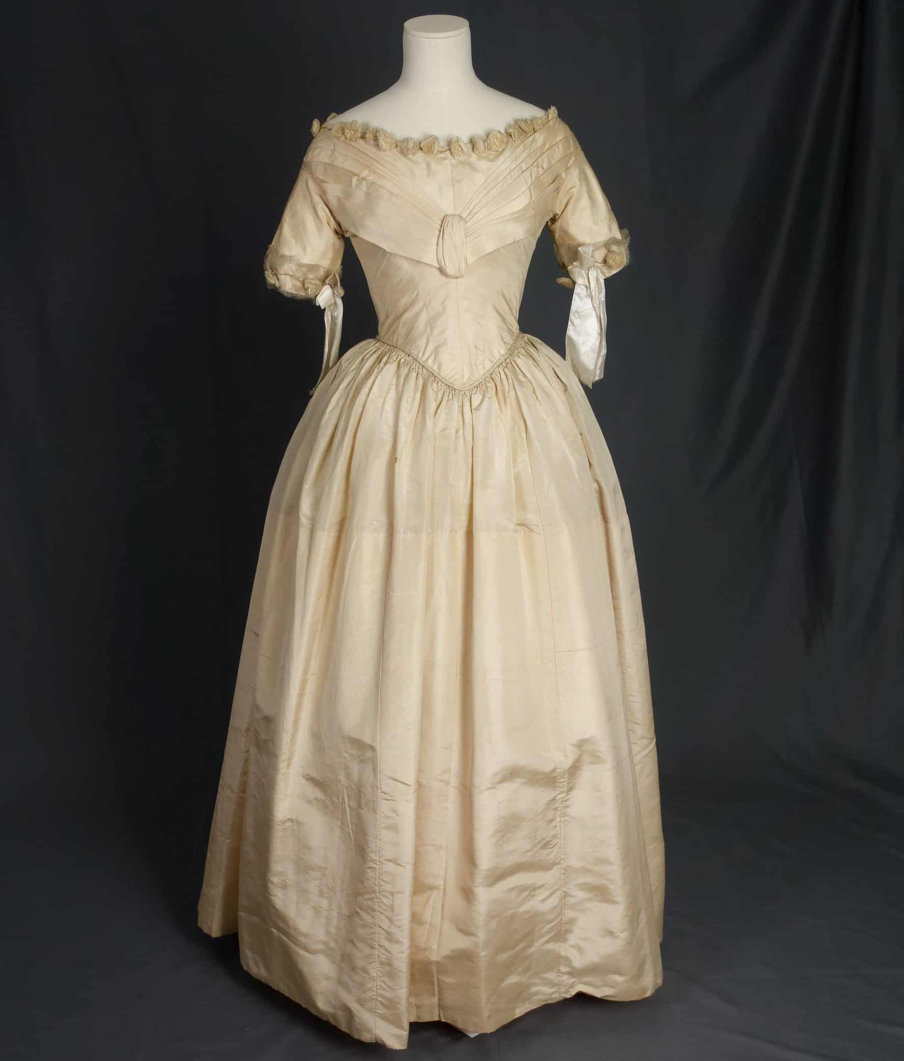 The wedding dress is styled on a mannequin. Made of cream colored ribbed silk. The bodice has a dropped shoulder line and a wide neckline and a pointed waist seam. The bodice has a V shaped pleating of fabric over the bust. The neckline is embellished with a row of fabric flowers. The short sleeves are straight and trimmed around the openings with tulle, more flowers, and silk ribbon bows. The skirt is gathered into the pointed waist all around. Gathered across the front and cartridge pleated across the back. The center back opening extends for 22" and fastens with hooks and eyes in the bodice. The bodice is lined with glazed white cotton.