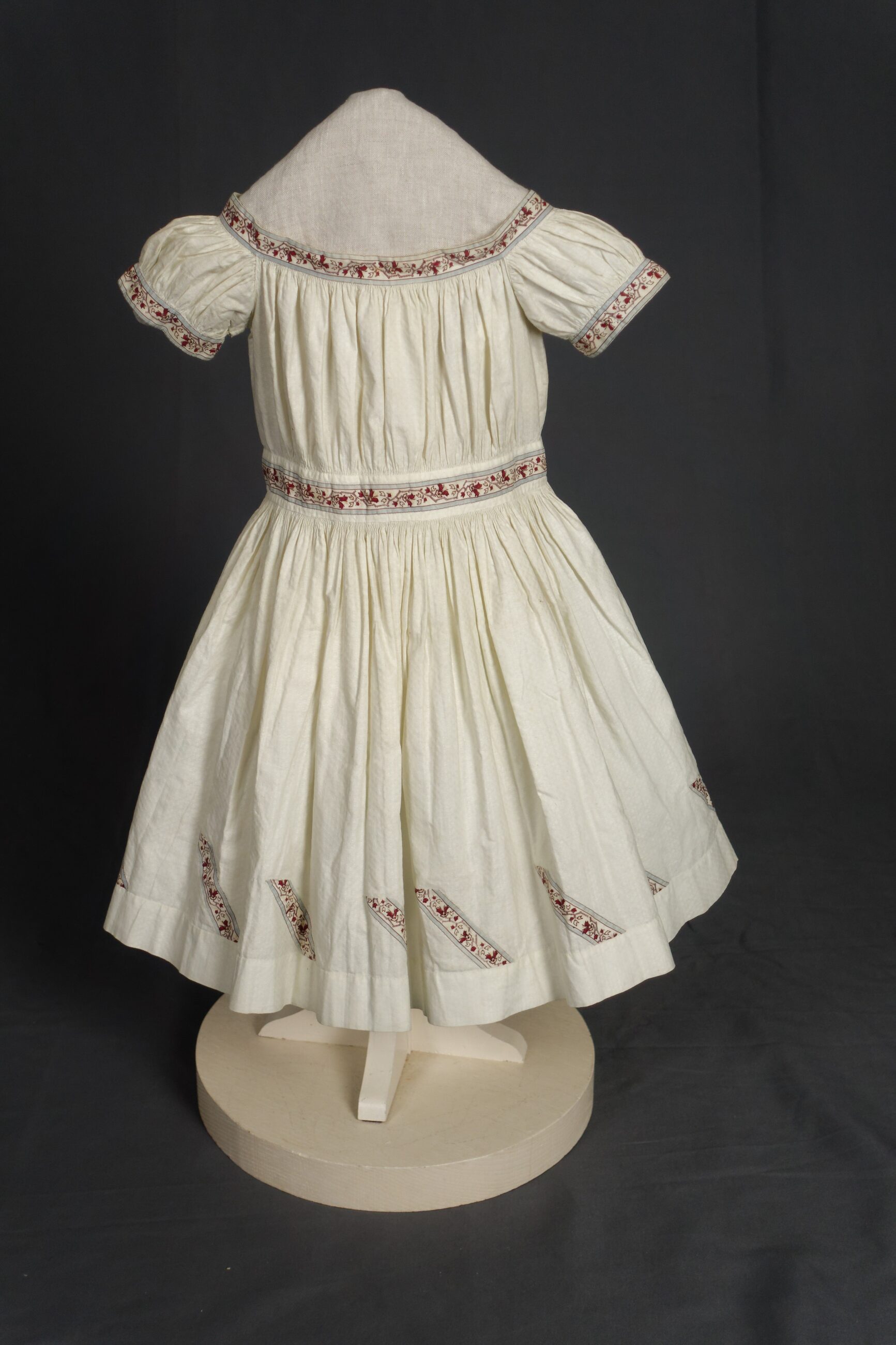An ivory-colored children's dress displayed on a mannequin stand against a dark grey background. The bodice has Short, puffed sleeves and a gathered waistline. Fine red and brown floral border trim accent the collar edges, sleeves, and waistband. Angled, designs featuring matching floral borders line the bottom hem.