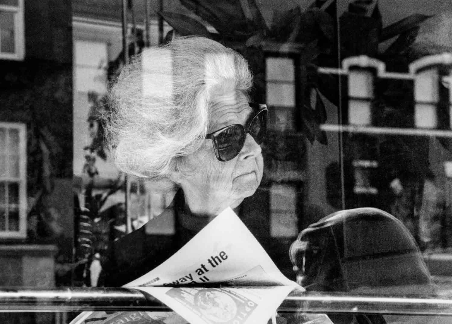 Photograph of a matured woman in the window of a store or restauraunt. She has a book or magazine open in her hands and is looking off to the side at something. She has on sunglasses and her white hair is in an updo.