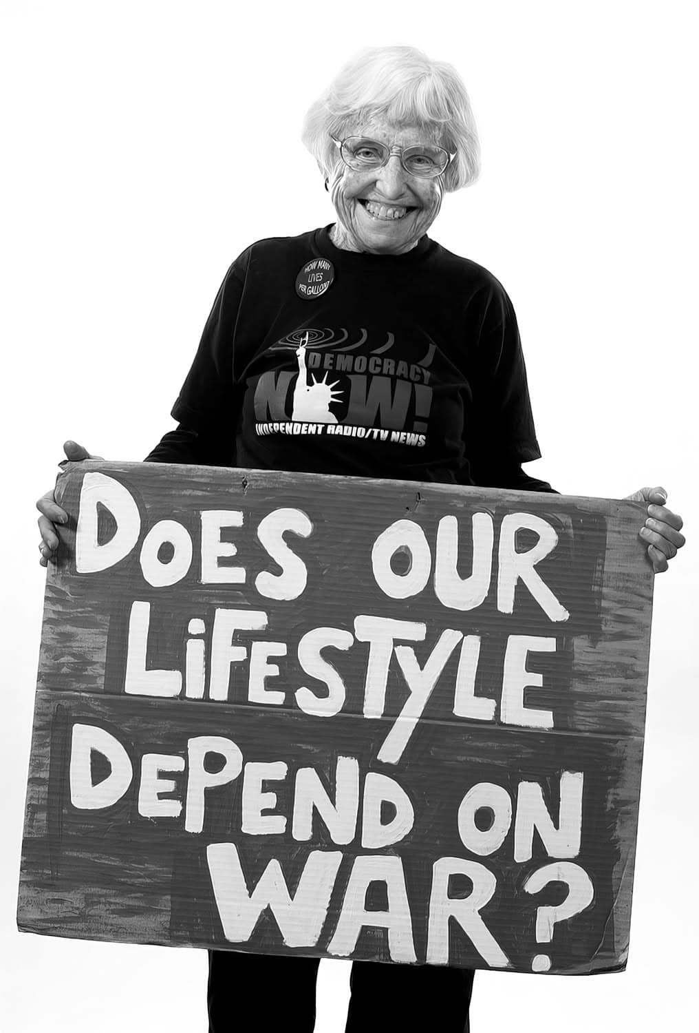 Black-and-white photograph of a matured woman with short white hair and round glasses smiling at the camera. She is wearing a Democracy Now t-shirt and holding a large sign that reads: "Does our Lifestyle Depend on WAR?"