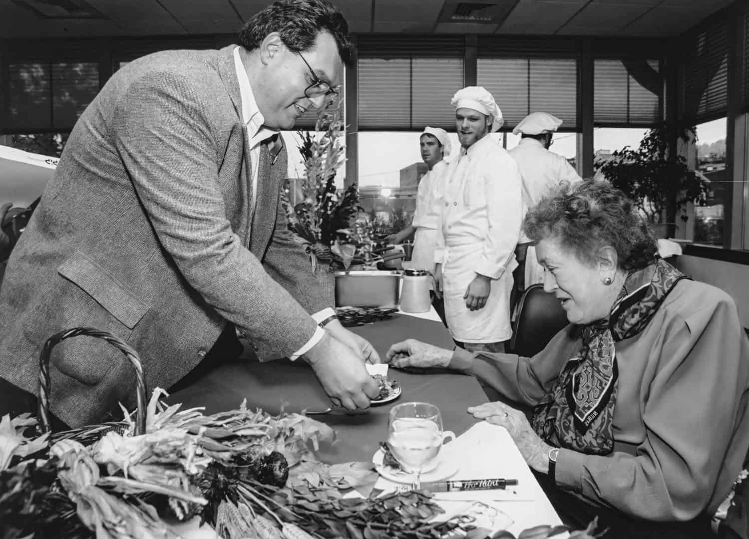 Photograph of man and a middle aged Julia Child. The man is standing across the table from Julia Child and handing her something. She is looking at what he is handing to her. Three men in white chef uniforms can be seen in the background.