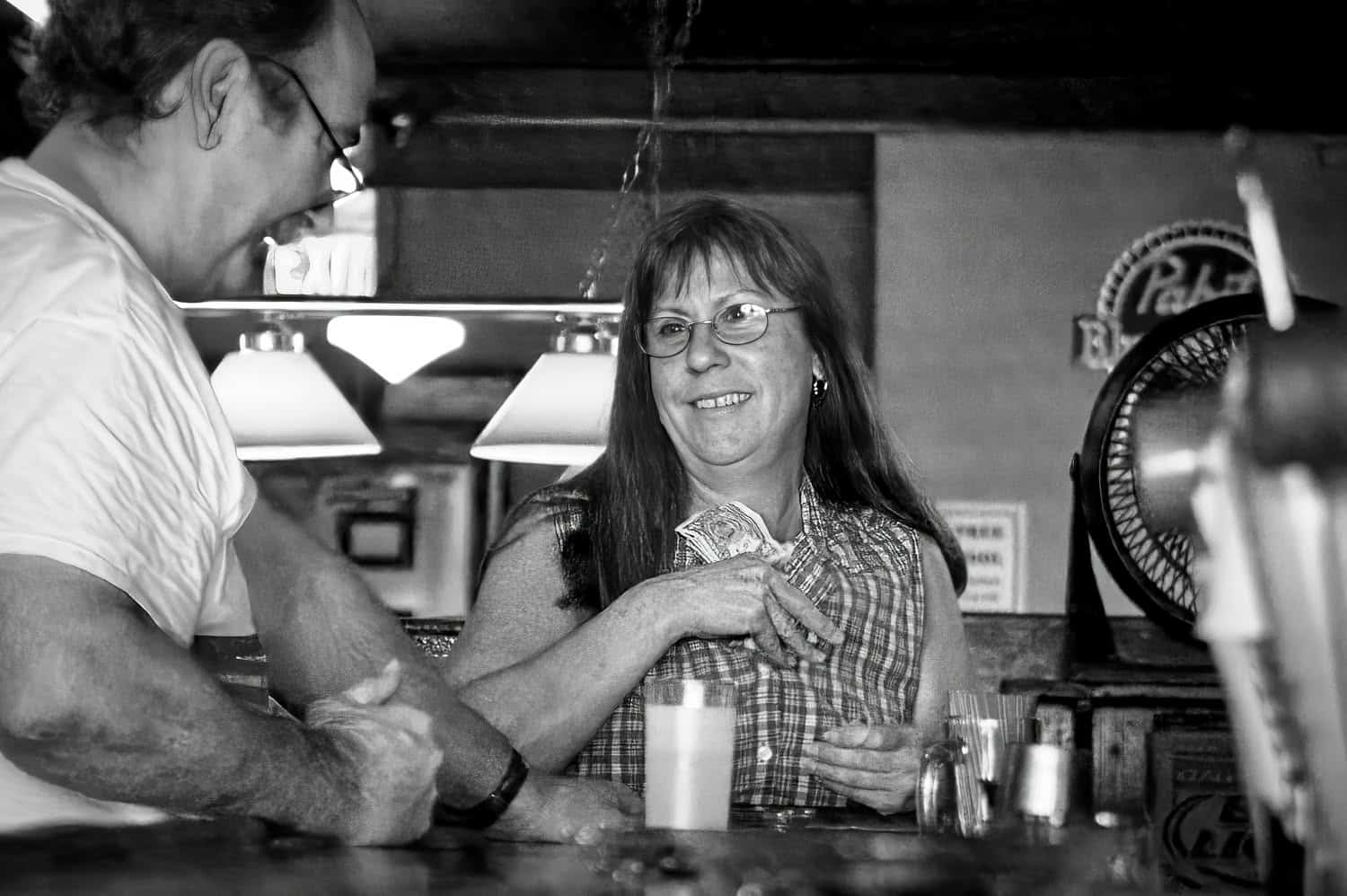 Black-and-white photograph of a middle aged woman with dark brown hair with banks. She is wearing glasses and smiling at the man next to her. He is looking down at the table.