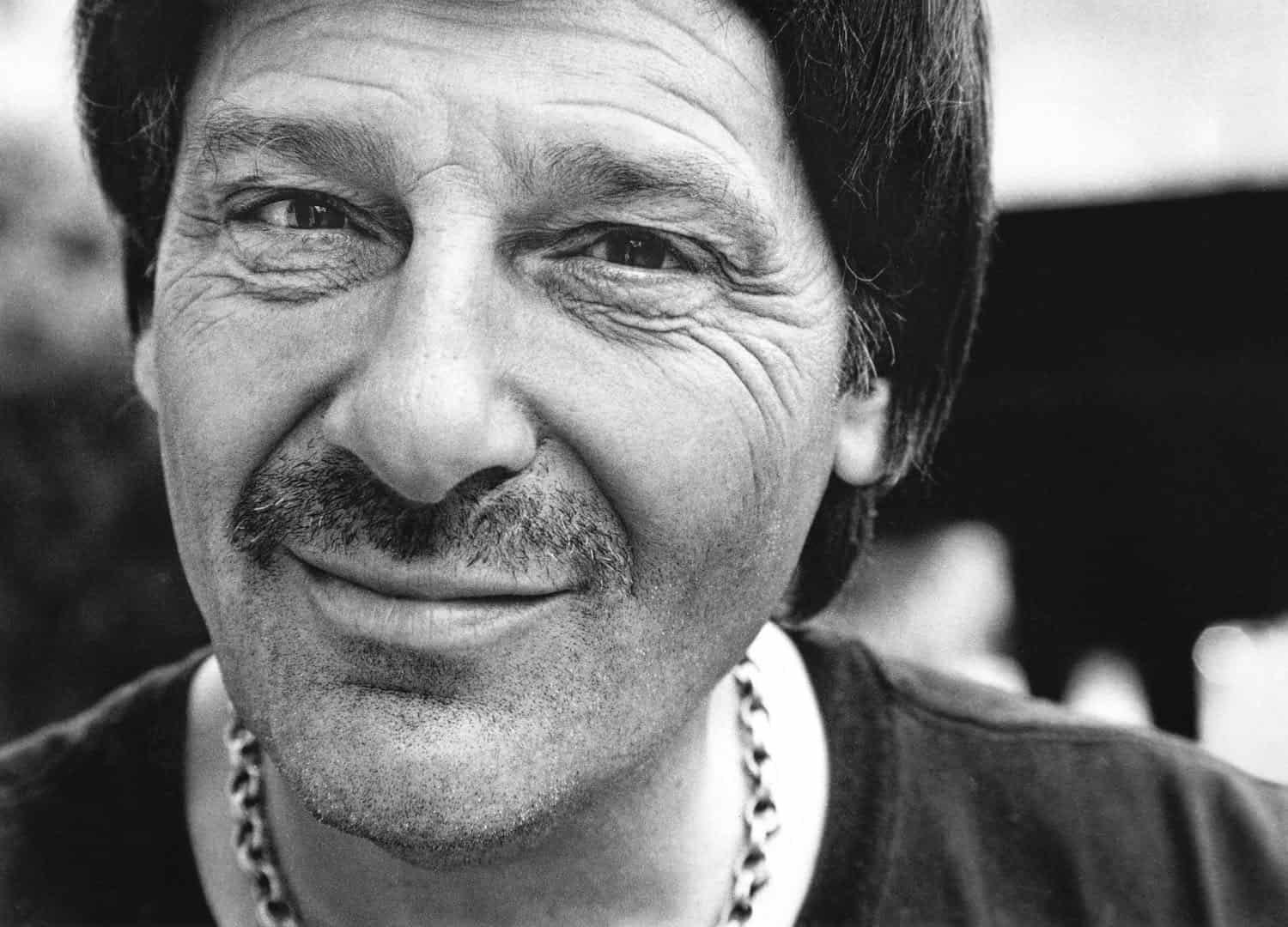 Black-and-white portrait photograph of a middle aged man with a scruffy beard and mustache. He has full dark hair and is smirking at the camera.