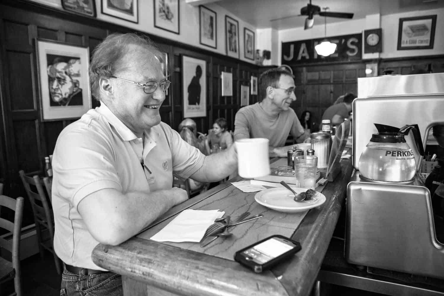 Black-and-white photograph of two men sitting at the bar at Jake's Diner. The man closest to the camera is looking behind the counter and smiling. He is holding a coffee. The man next to him is facing toward the counter as well. Framed photographs line the walls and a large sign saying "JAKES" is above a door.