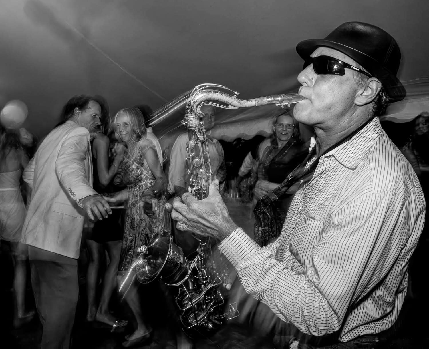 Black-and-white photograph of a matured man in a button-down shirt playing the saxophone. He also has on a black bat and sunglasses. People are dancing in the background.